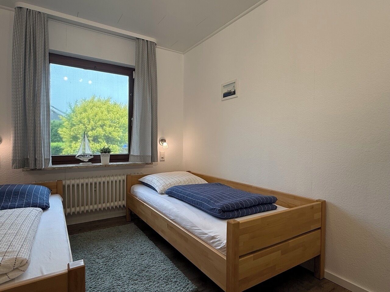 Une petite pièce propre et bien rangée, avec un lit simple, une fenêtre donnant sur un espace verdoyant, un radiateur et une petite table avec une lampe.