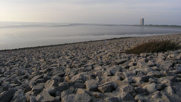 A rocky beach with a calm sea and a distant building. - FeWo Seestern... at home on vacation (Büsumer Deichhausen)