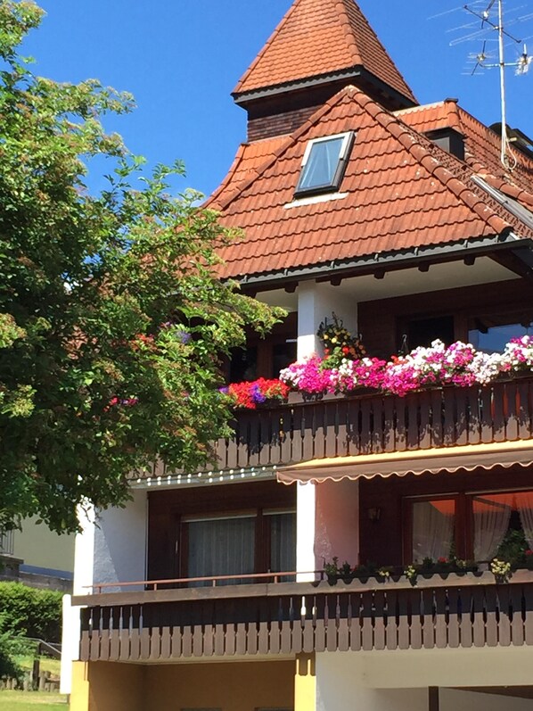 Un edificio de dos pisos con techo de tejas rojas, un balcón con jardineras y una ventana pequeña en el piso superior.