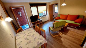 Una sala de estar acogedora con piso de madera, un sofá verde con cojines naranjas, una mesa de comedor con sillas y un televisor.
