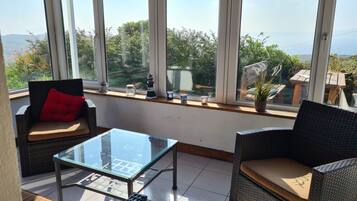 A sunlit room with wicker furniture, a glass table, and a view of greenery and hills.