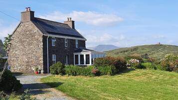 A stone house with a solar panel roof, a covered porch, and a grassy yard with a hill in the background.