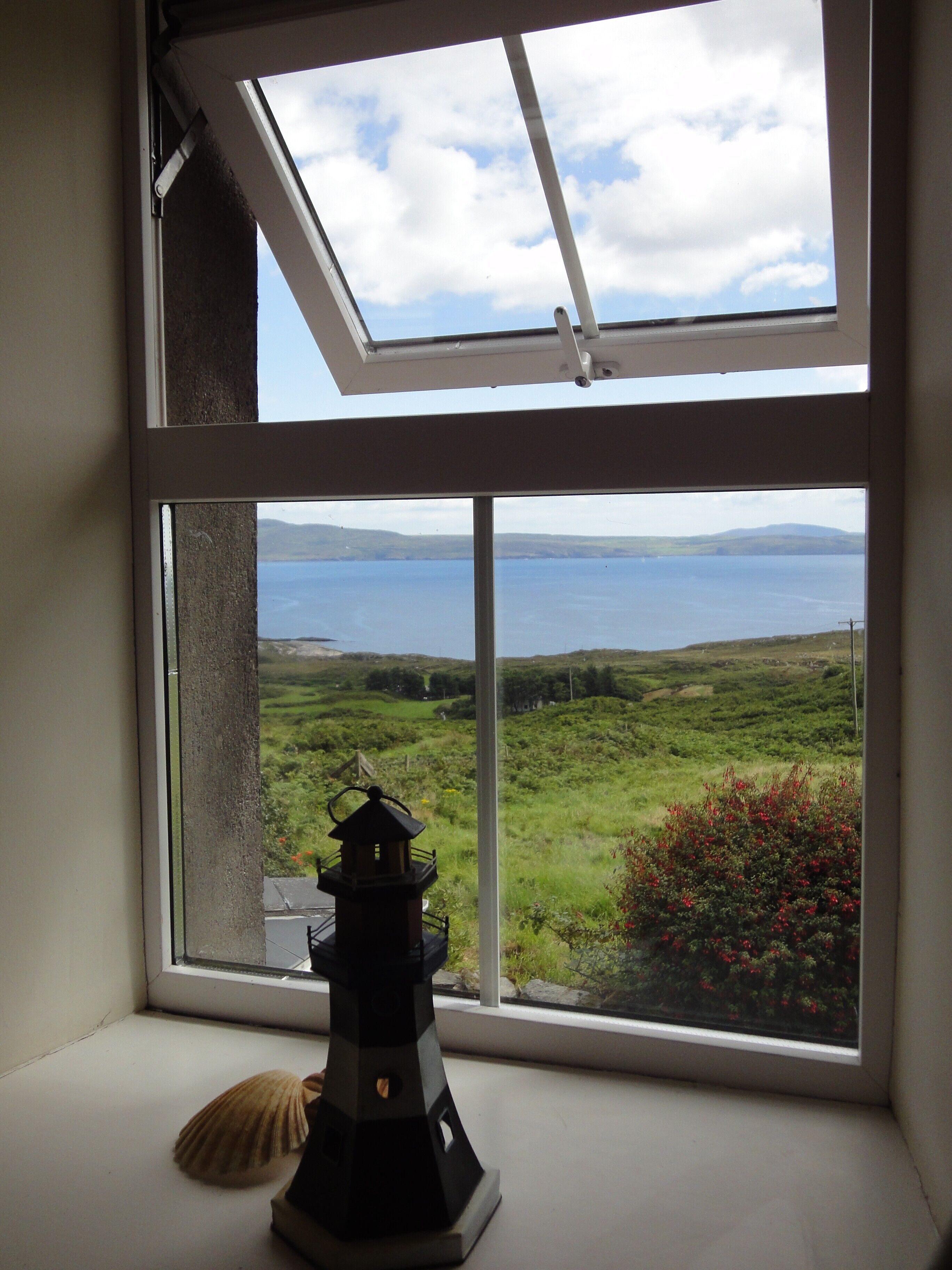 A window with a view of a coastal landscape, a lighthouse model, and a seashell.
