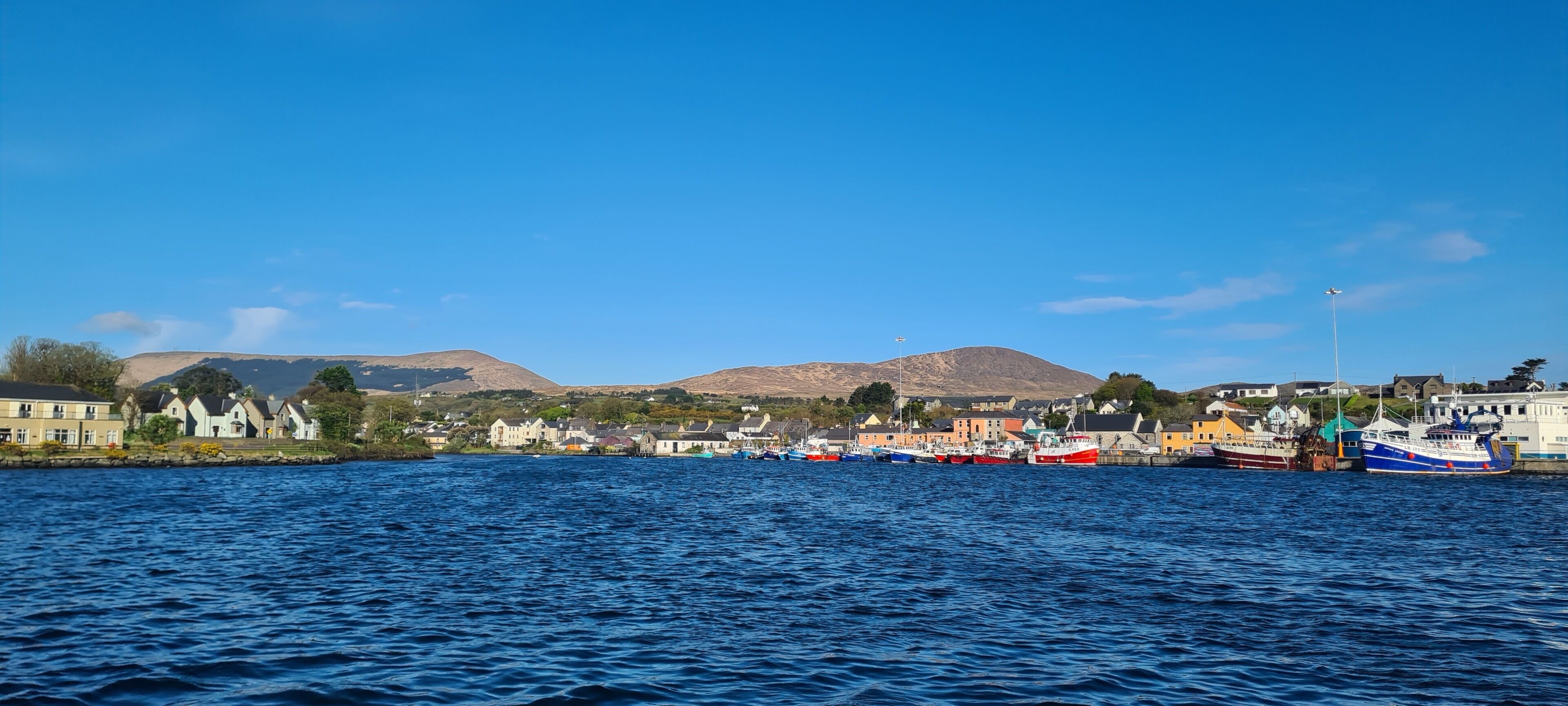 A harbor with boats docked, buildings along the shore, and hills in the background.