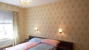 Une chambre à coucher avec un lit, deux lampes de chevet, un lustre, un papier peint à motifs floraux et une fenêtre avec des rideaux.