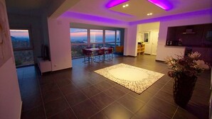 A modern living room with a large window, a dining area, and a kitchen.