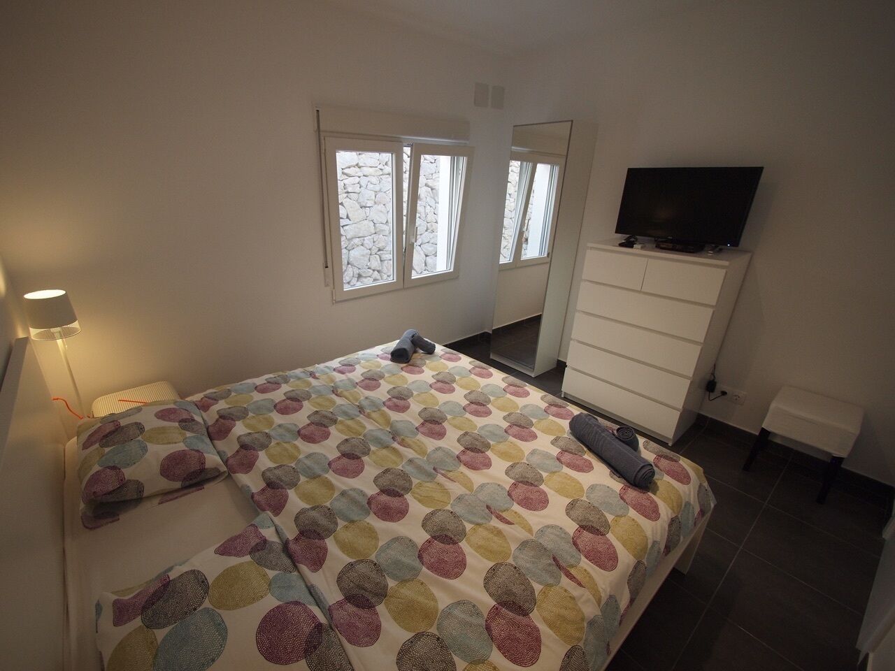 A bedroom with a bed, a TV on a dresser, and a window with a stone wall view.