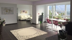 A modern living room with a white sofa, a dining area with red chairs, and a large window offering a view of the outdoors.