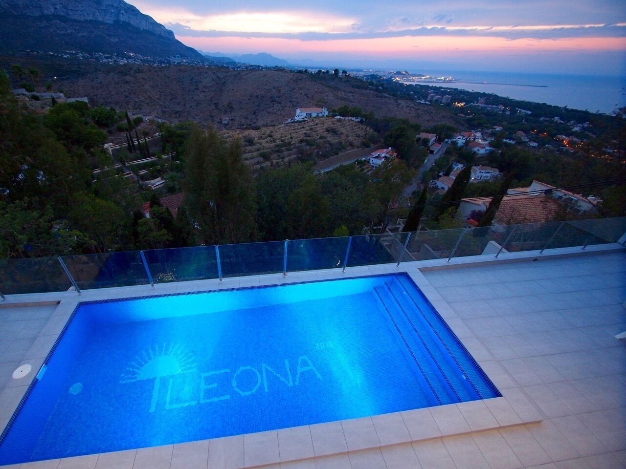 Una piscina con la palabra "TELEONA" en el agua, rodeada por un suelo de baldosas y barandilla de vidrio, con vistas a un paisaje montañoso y una masa de agua.