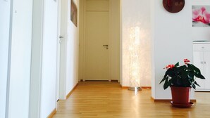 A hallway with wooden flooring, white walls, a door at the end, a wall-mounted light fixture, and a potted plant.