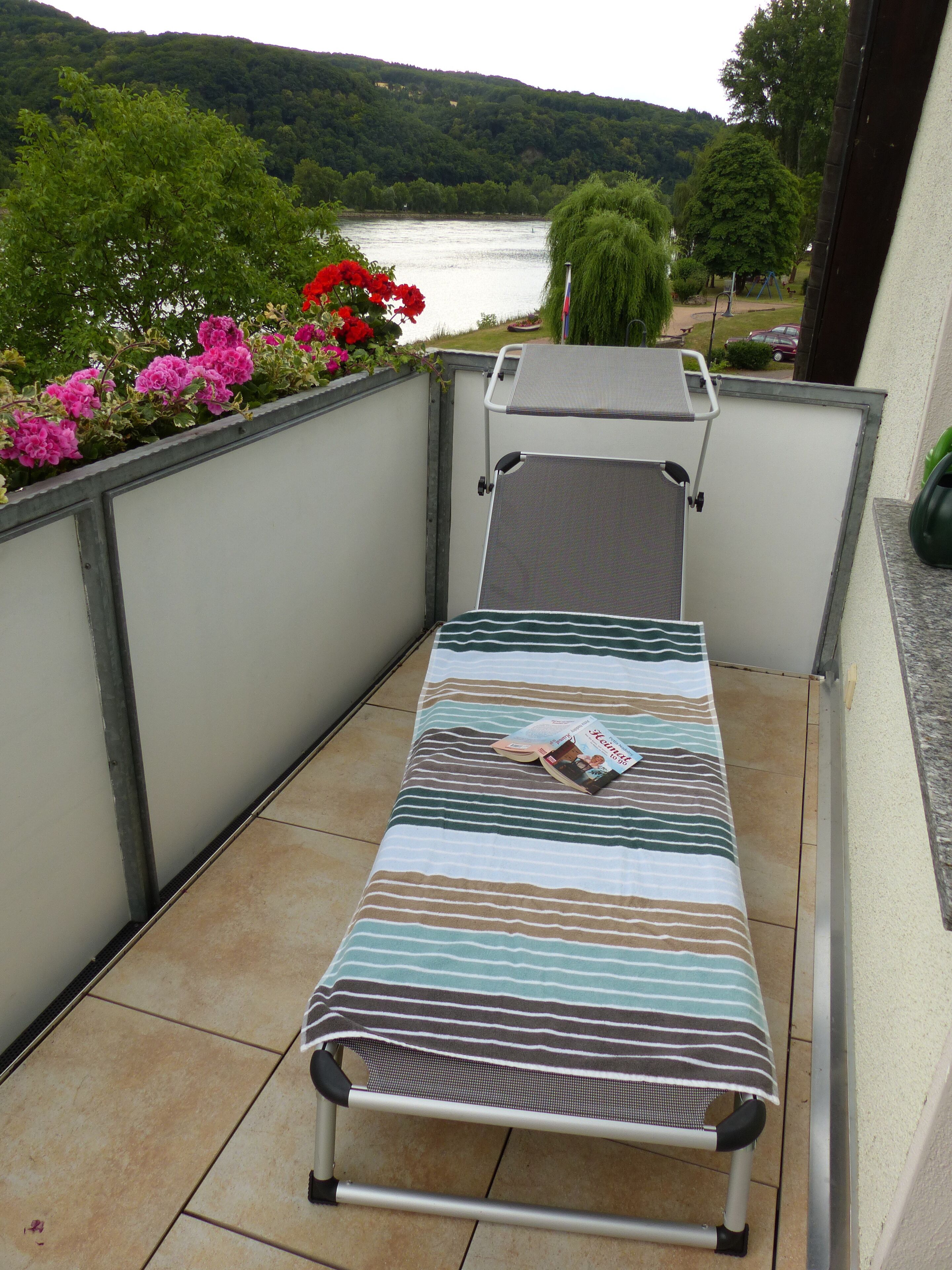 Un petit balcon avec un seul fauteuil, une vue sur un lac et un jardin parsemé de fleurs roses.