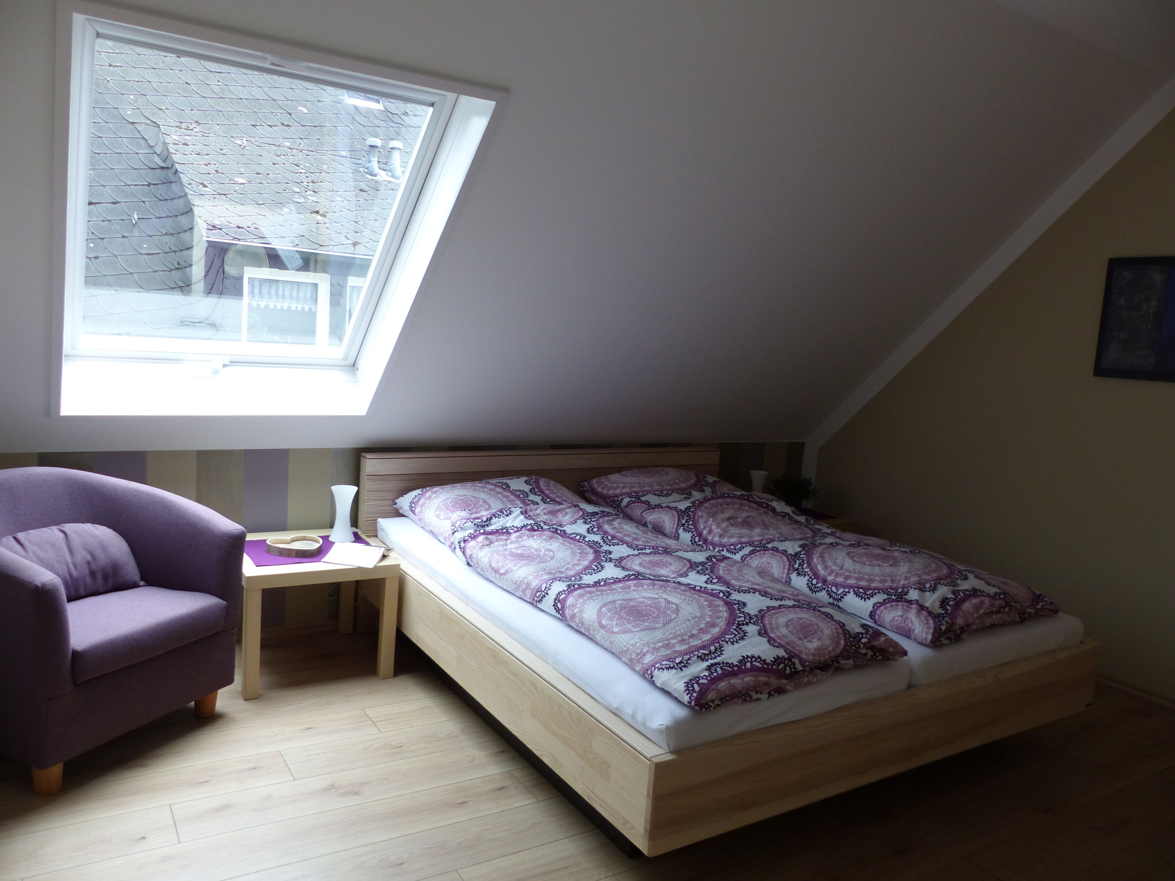 Une chambre avec un plafond en pente, un lit avec une literie à motifs, un fauteuil violet, une table de chevet avec une lampe et une fenêtre donnant sur la rue.