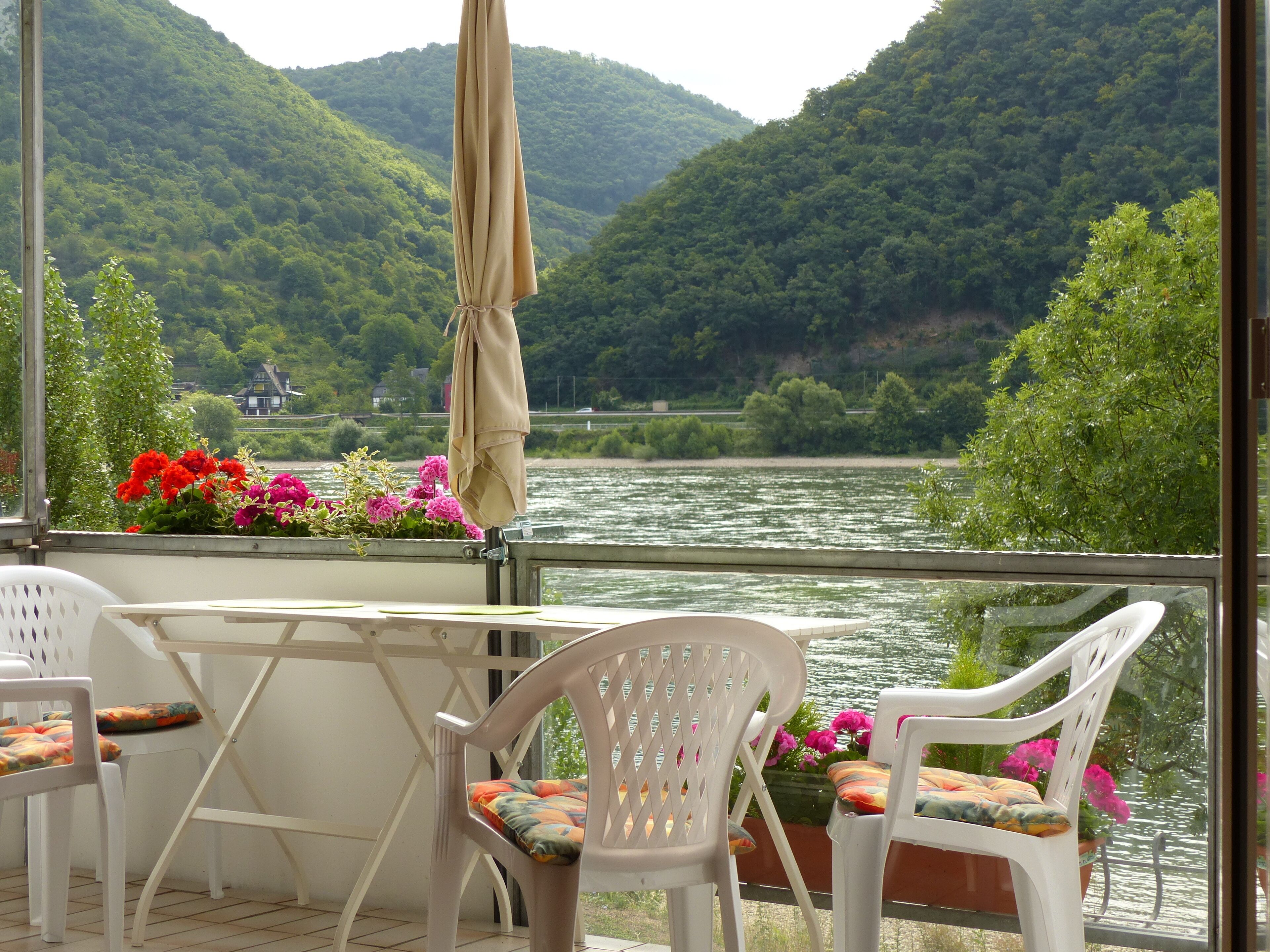 A balcony with a table and chairs, overlooking a river and forested hills.