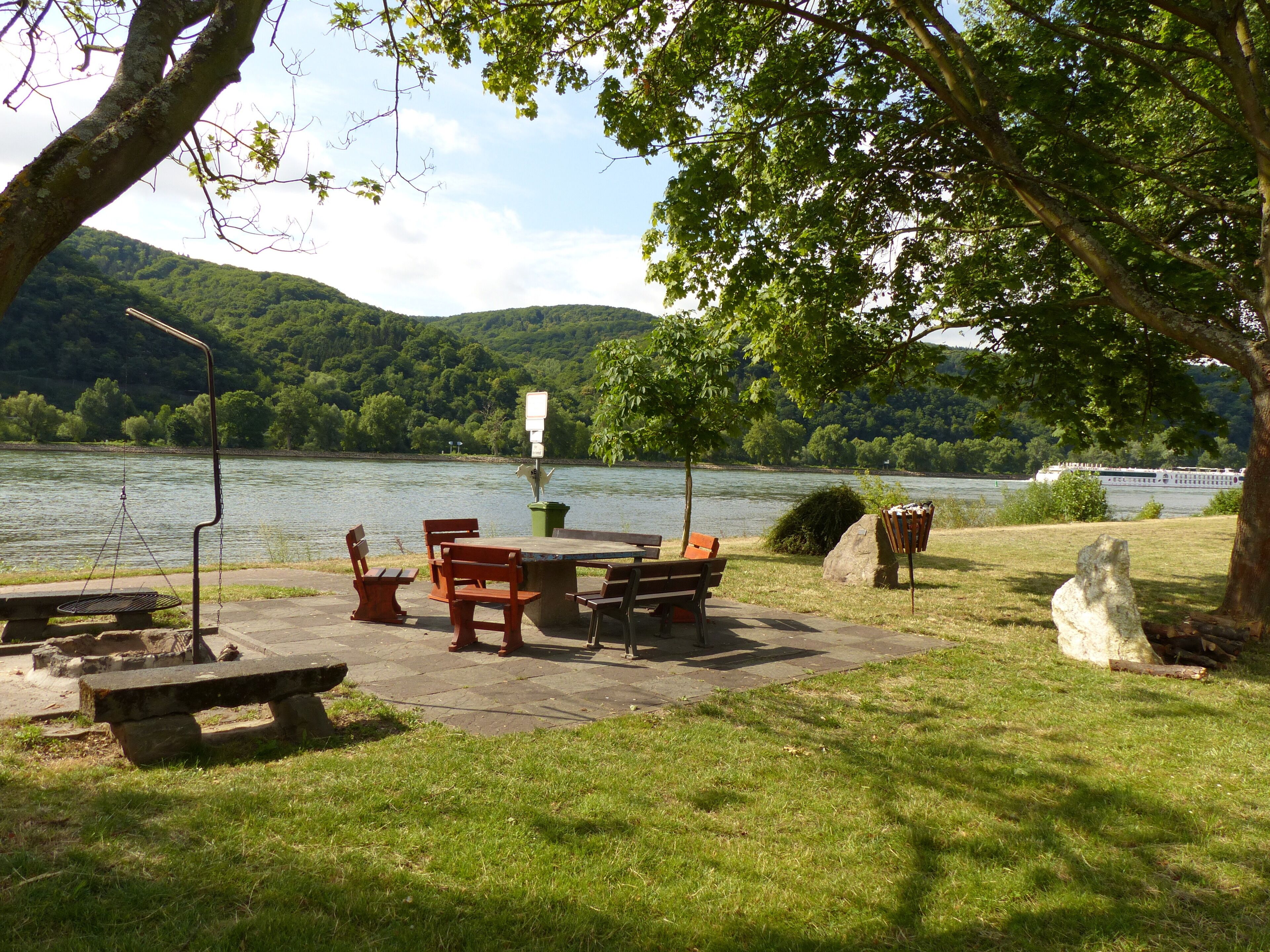 Ein Picknickplatz am See mit Picknicktischen und Bänken, umgeben von Bäumen und Blick auf einen Fluss und Hügel.