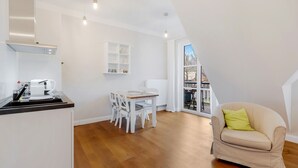 Et moderne køkken med hvide skabe, et spisebord med stole, en beige sofa med en grøn pude, og et stort vindue med udsigt udad.
