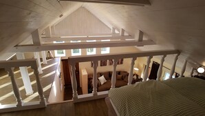 A high-ceilinged attic bedroom with a sloped roof, wooden beams, and a view of the lower level. - Summer in the old stable (Iserlohn)