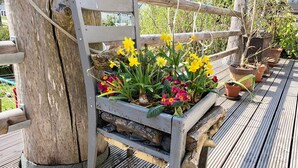 Une terrasse en bois avec un banc en métal sur lequel est posé un jardinier contenant des fleurs jaunes, rouges et roses.
