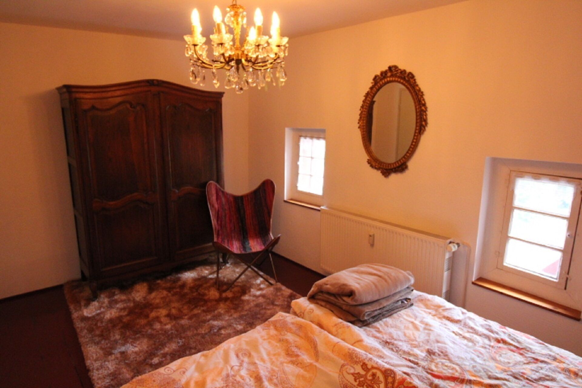 A bedroom with a large wooden wardrobe, a single chair, a bed with a patterned bedspread, a chandelier, and a mirror on the wall.