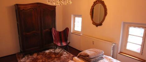 A bedroom with a large wooden wardrobe, a single chair, a bed with a patterned bedspread, a chandelier, and a mirror on the wall.