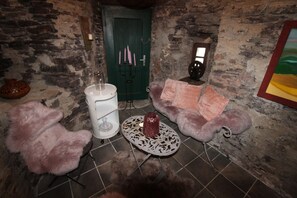A rustic room with a stone wall, a green door, a white barrel, a patterned table, and two fluffy chairs.