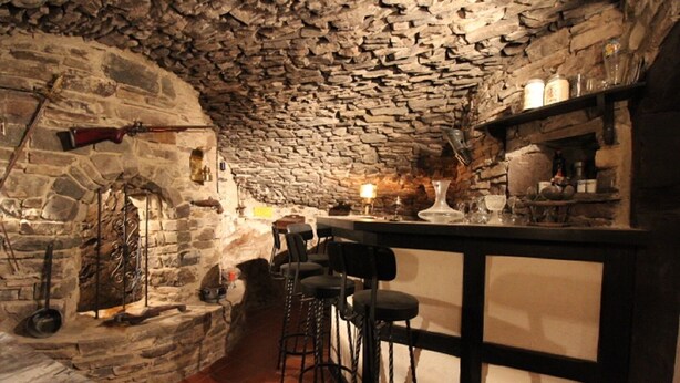 Eine Bar mit steinernen Wänden und rustikalem Ambiente, inklusive Kamin, Barhocker und einer Ablage mit diversen Gegenständen.