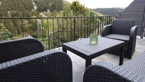 Ein Balkon mit Korbmöbeln und einem Tisch, mit Blick auf ein bewaldetes Tal.