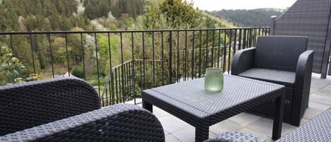 A balcony with wicker furniture and a table, overlooking a forested valley.
