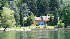 Una casa junto al lago con fachada de piedra, ventanas amplias y un patio visible.