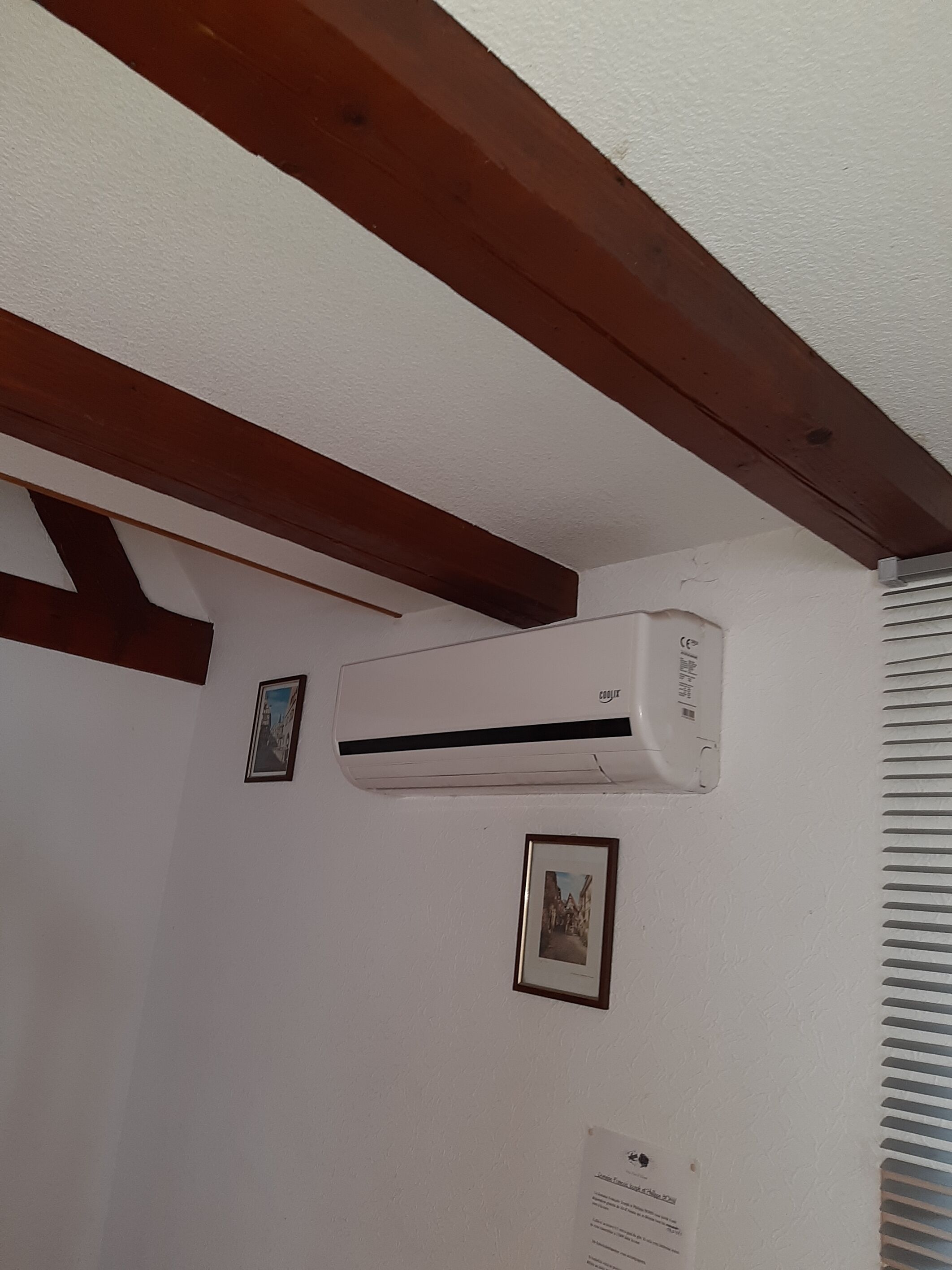 Ein Zimmer mit weißer Decke, Holzbalken, einer Klimaanlage und zwei gerahmten Bildern an der Wand.