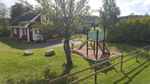 Un parque infantil con tobogán y estructuras para escalar, rodeado de césped y un edificio rojo.