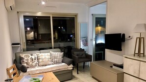A modern living room with a sofa, a dining table, a TV, and a kitchen area.