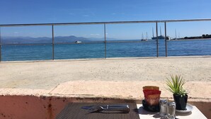 A table with a metal frame, salt and pepper shakers, a small potted plant, and a bag of snacks on a beach overlooking a marina with boats and distant mountains.