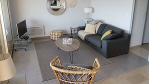 A modern living room with a grey sofa, a round coffee table, a wicker chair, a glass TV stand, and a patterned rug. - 4-room apartment 90 m² + Terrace 17 m². Sea Front, Panoramic Sea View (Bandol)