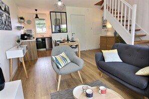 A modern living room with a grey sofa, a grey armchair, and a coffee table with mugs. A kitchen with a dining area is visible in the background. - Quiet house "Le Green" in the Domaine du Golf with swimming pool - near beaches (Pléneuf-Val-André)