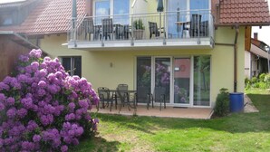 Ein zweistöckiges Haus mit einem Balkon, einer Terrasse und Gartenmöbeln sowie ein großer Strauch mit lila Blüten.