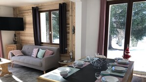 Une pièce chaleureuse avec un mur en bois, un canapé gris, une table à manger dressée avec des assiettes et des verres, et un téléviseur fixé au mur.