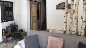Un salon avec un canapé gris, des coussins à motifs, une porte en bois, un miroir et des troncs de bouleaux.