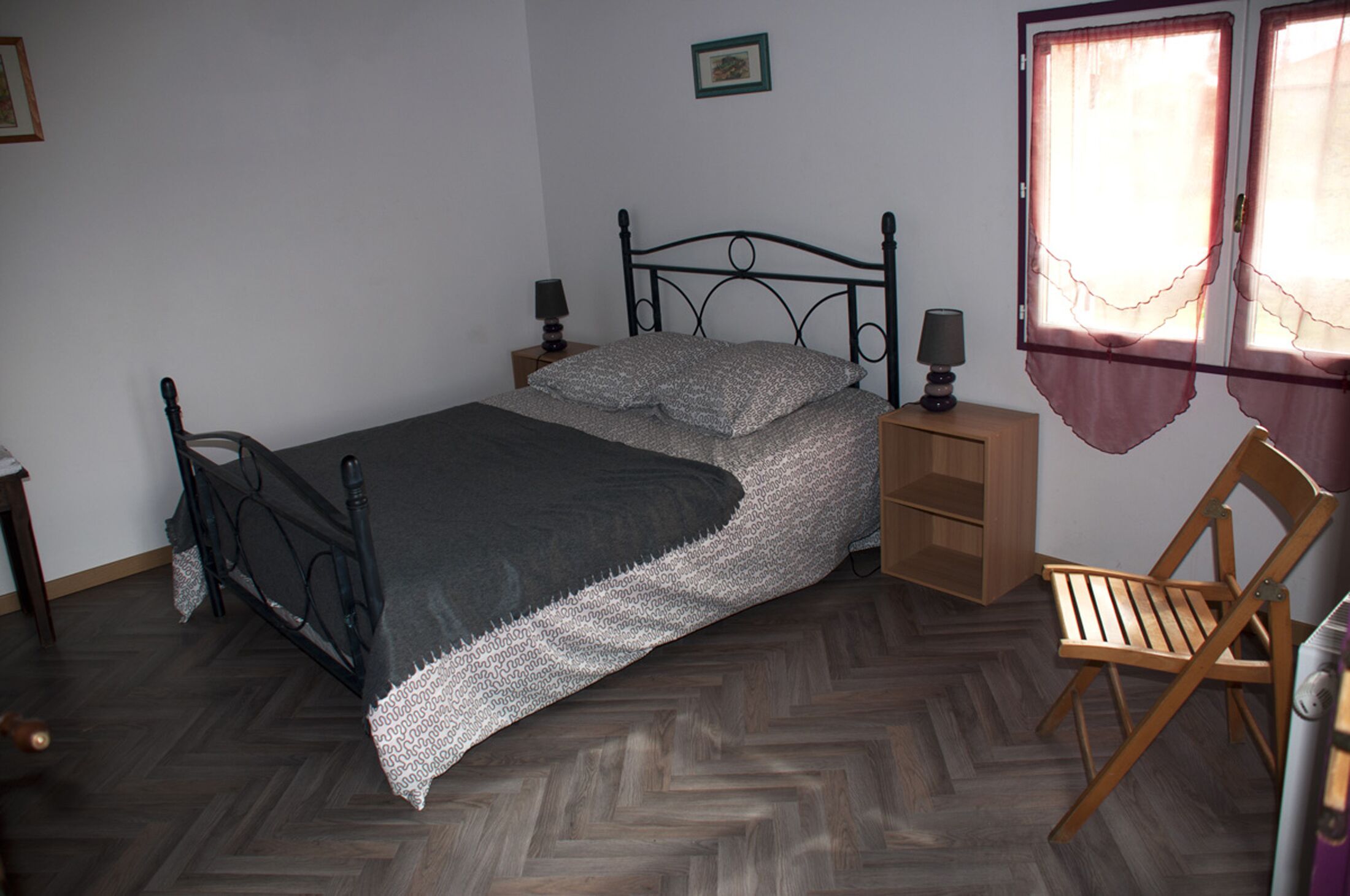 Une chambre à coucher avec un lit en métal, une chaise en bois, une table de chevet avec une lampe et une fenêtre avec des rideaux.