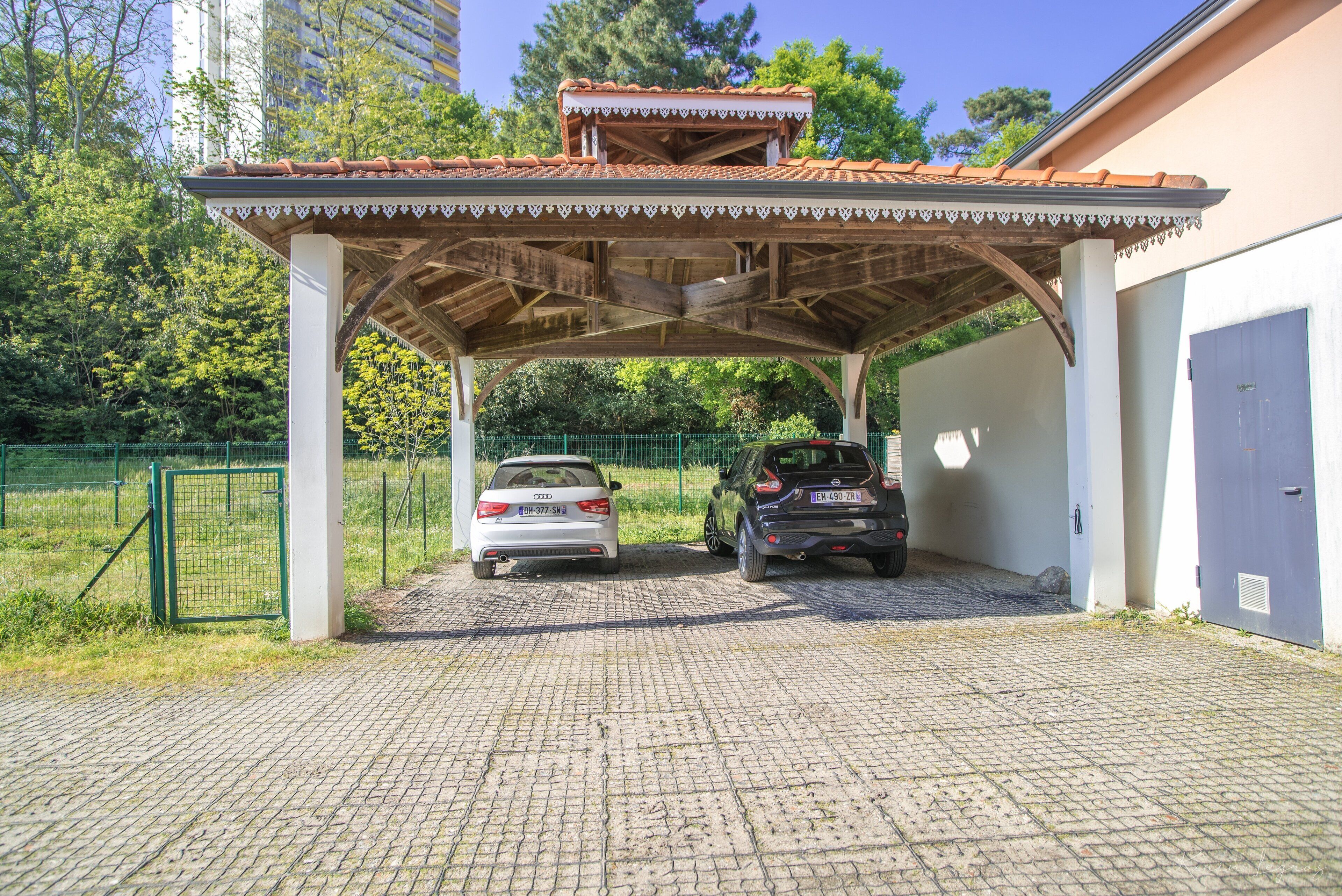 Un espace de stationnement couvert où sont garées deux voitures, un jardin clos et, au fond, un bâtiment avec un toit en tuiles.