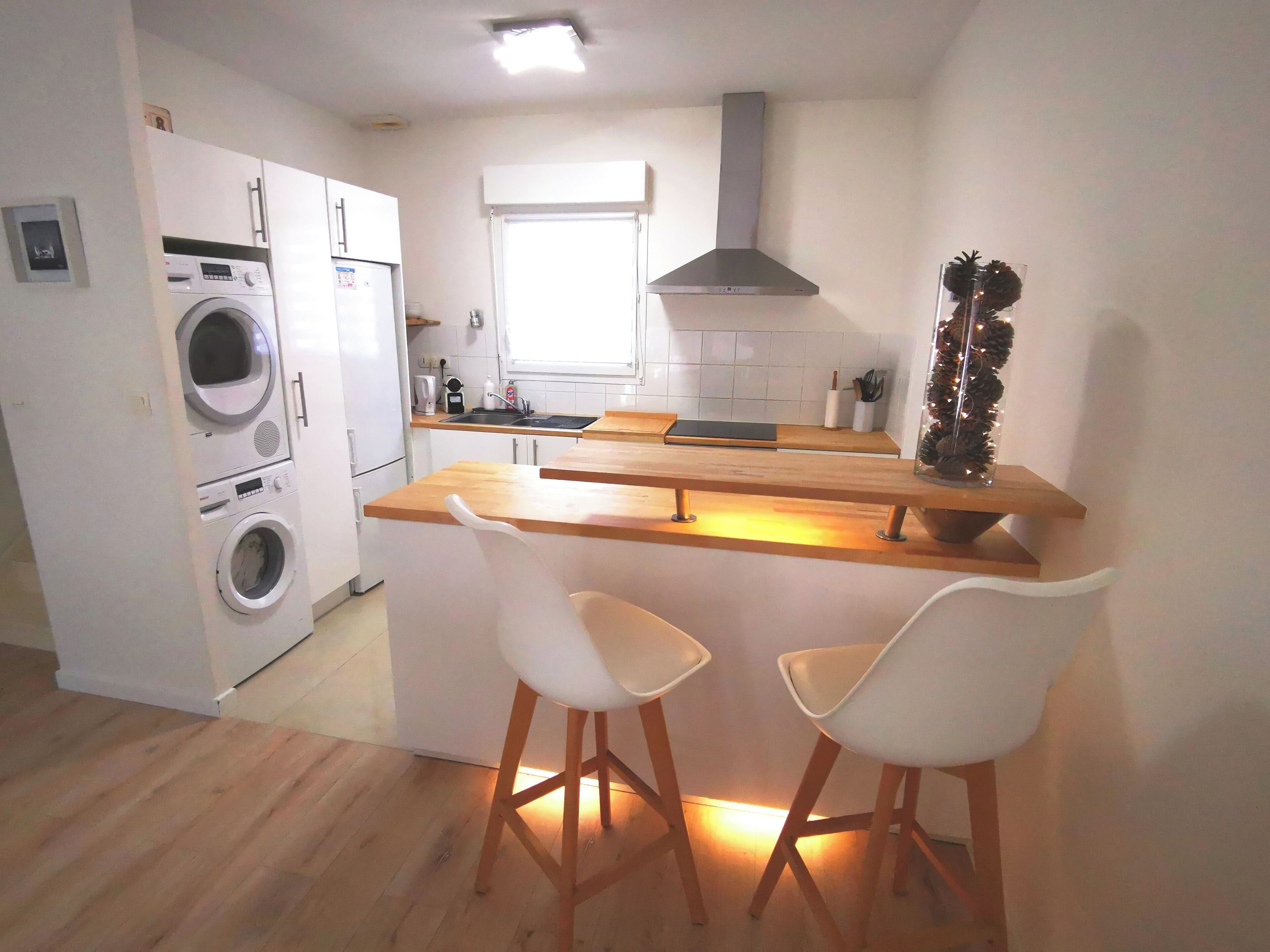 Una cocina compacta con lavadora, estufa y lavabo. Hay dos taburetes blancos y una encimera de madera.