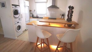 Une cuisine compacte équipée d’un lave-linge, d’une cuisinière et d’un évier. On y trouve deux tabourets de bar blancs et un comptoir en bois.
