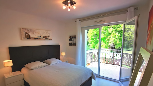 Une chambre à coucher comprenant un lit, des lampes de chevet, une table de chevet, une chaise et une grande porte-fenêtre coulissante donnant sur un balcon extérieur.