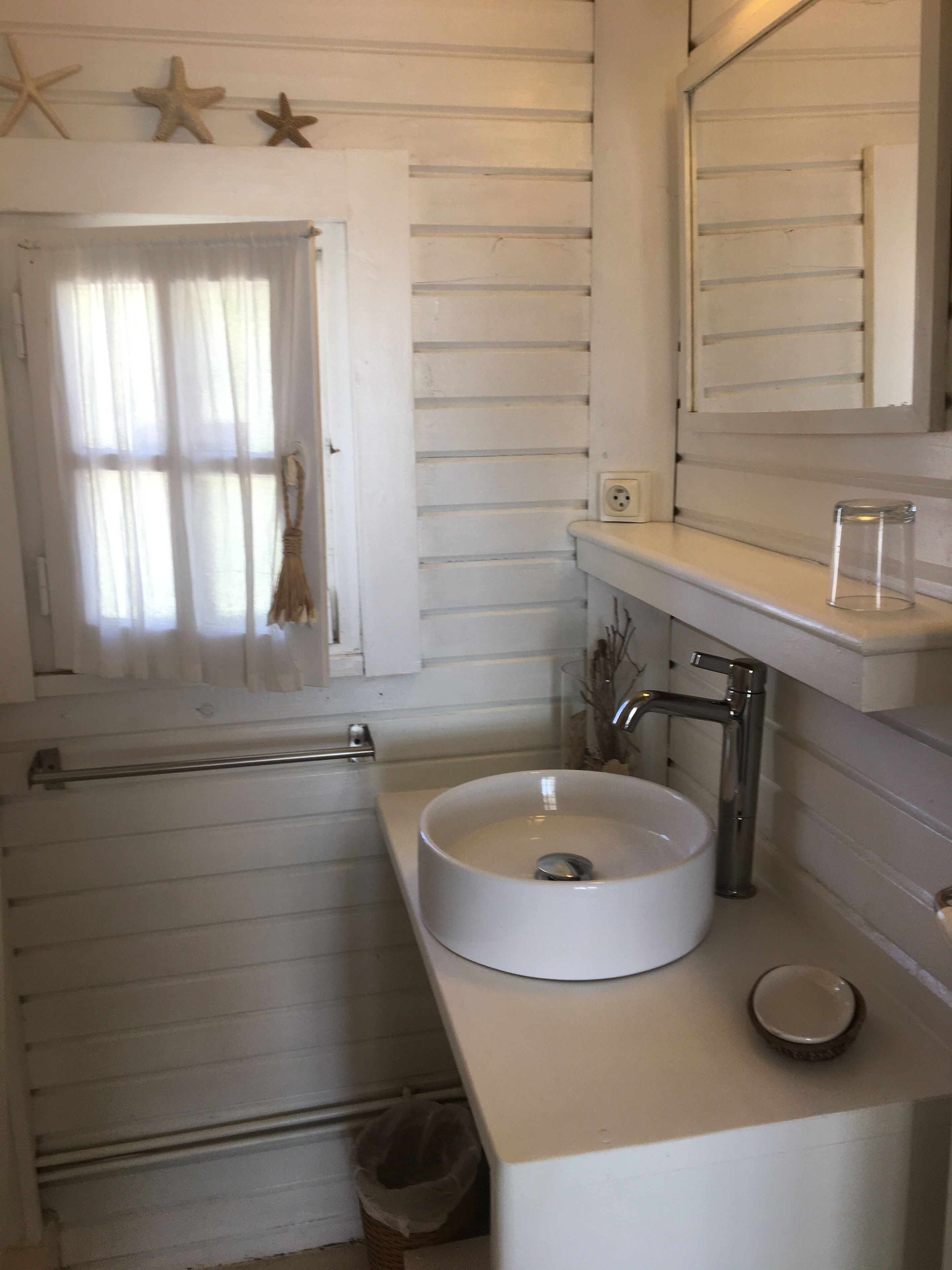 A compact bathroom with a round sink, a mirror, and a window with curtains.