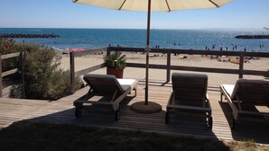 Um deck de madeira com cadeiras de descanso e um grande guarda-sol, com vista para uma praia com pessoas e um paredão rochoso.