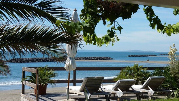Um espaço de relaxamento à beira-mar, com espreguiçadeiras de madeira e um guarda-sol branco, com vista para um mar calmo e formações rochosas.