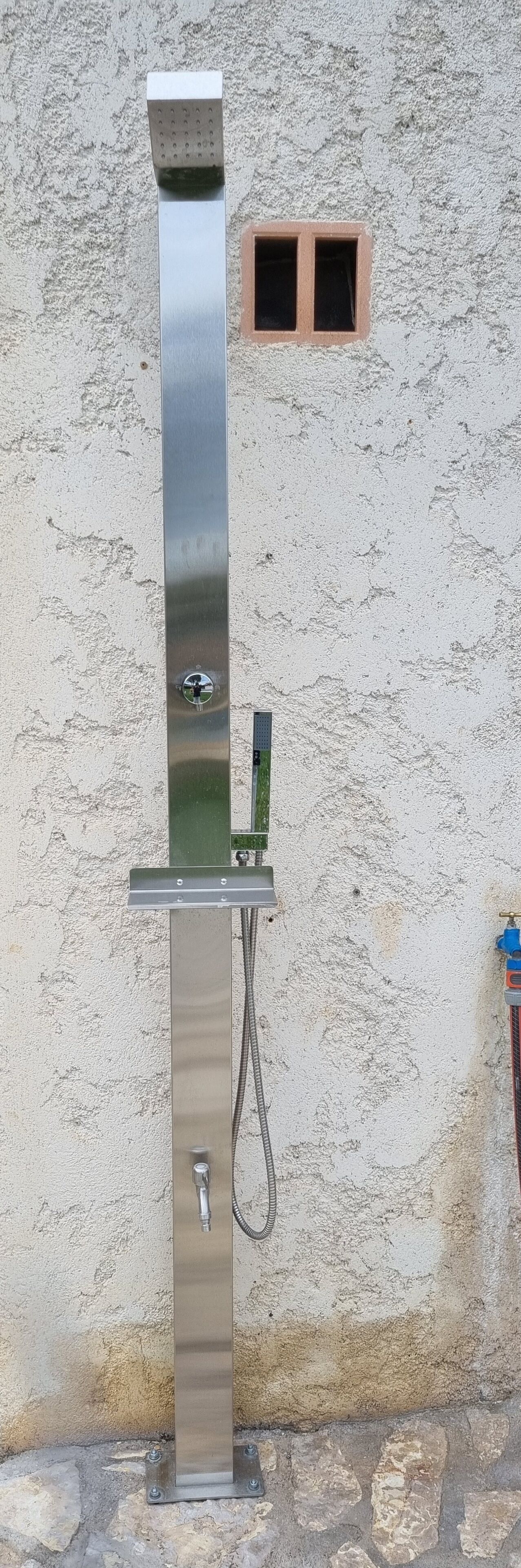 Una fuente de agua metálica exterior con manguera y panel de control, ubicada frente a una pared texturizada con un pequeño ventanal.