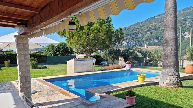 A pool area with stone tiles, a pergola with striped awning, and a view of a mountainous landscape.