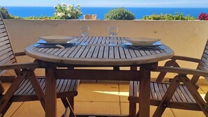Een terras buiten met een houten tafel en stoelen, uitzicht op de oceaan en een helderblauwe lucht.
