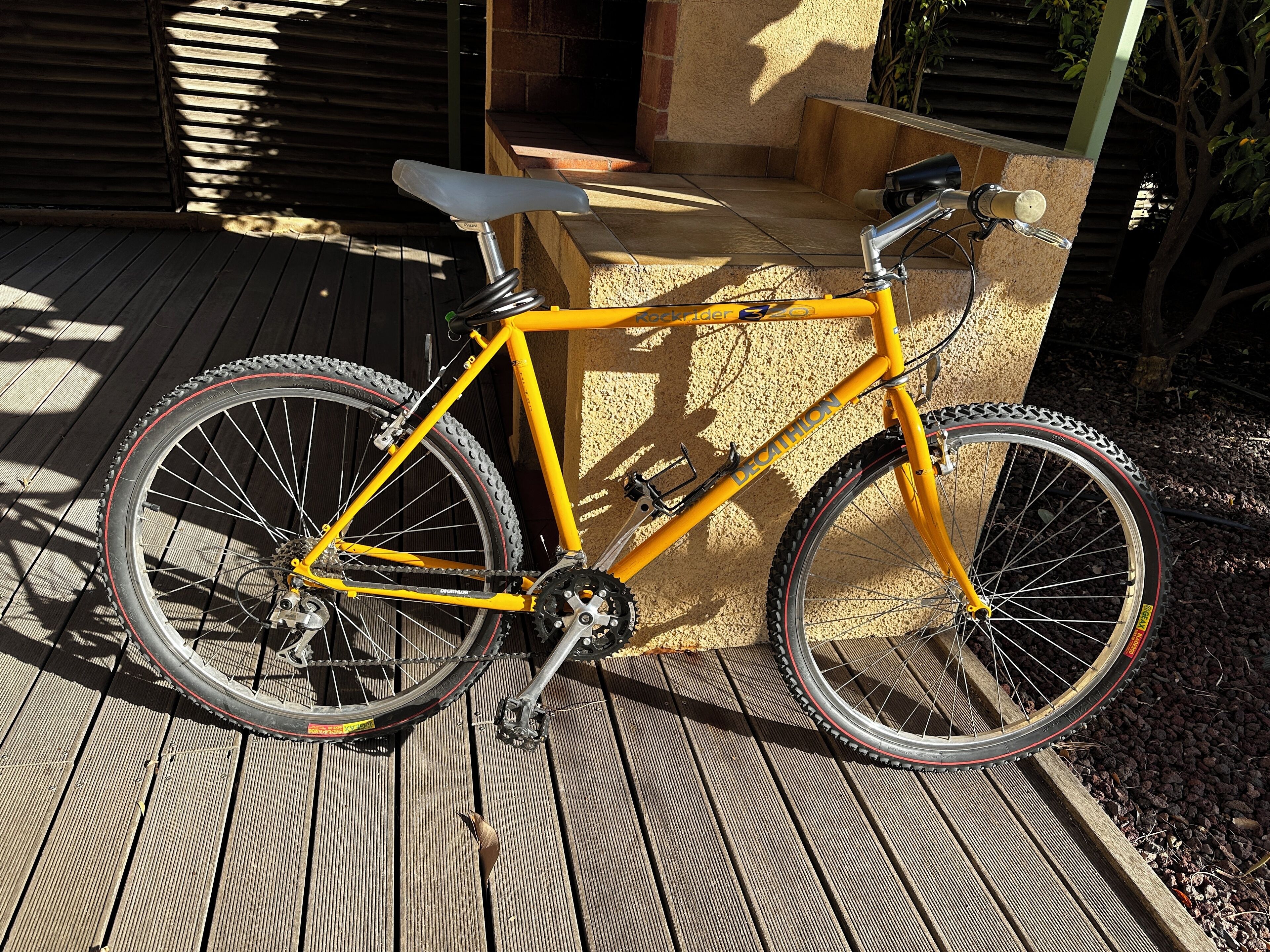 A yellow bicycle with a seat and handlebars, positioned on a wooden deck with a stone wall in the background.