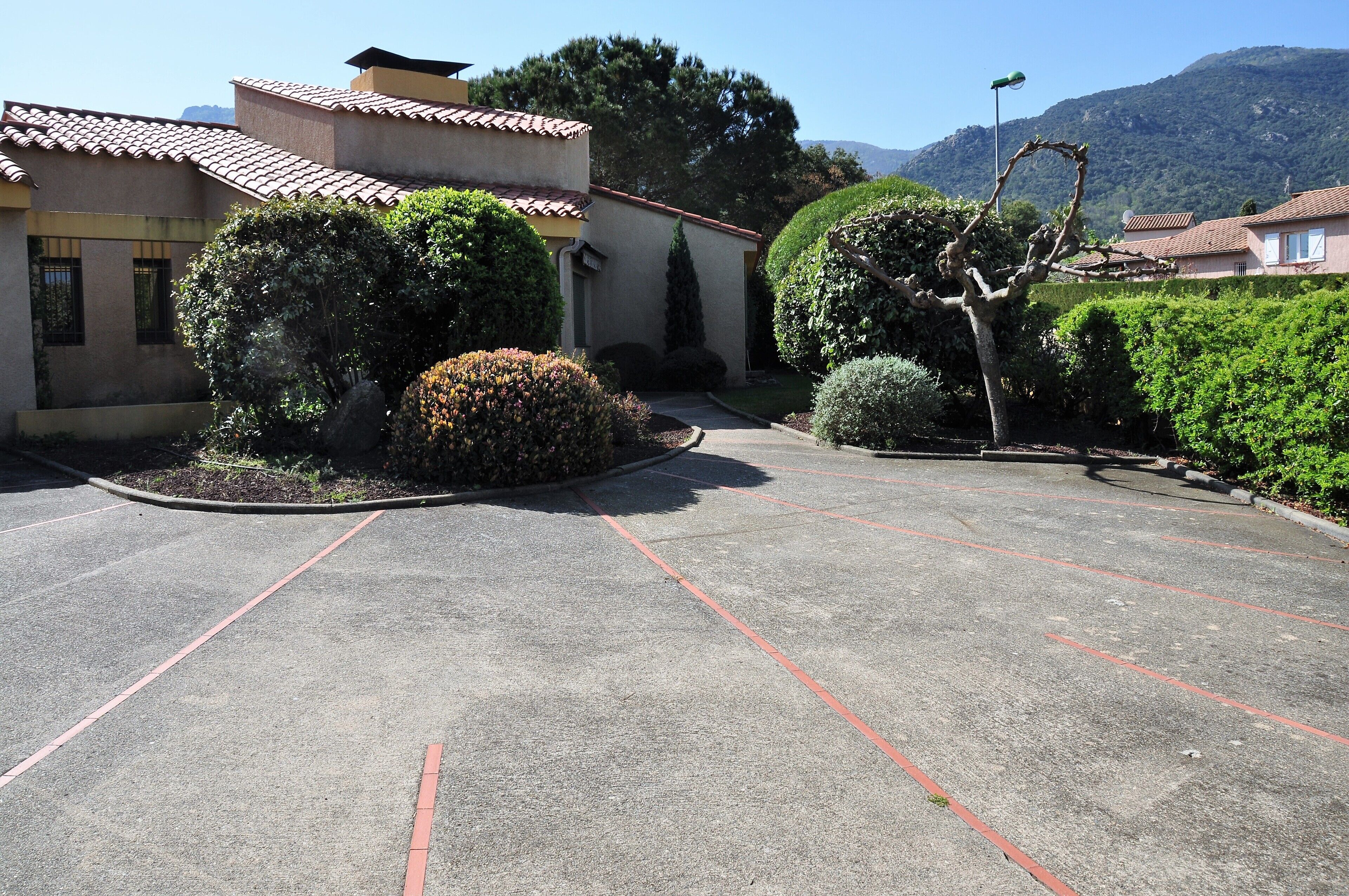 A parking lot with marked spaces, surrounded by well-maintained hedges and a building with a tiled roof.
